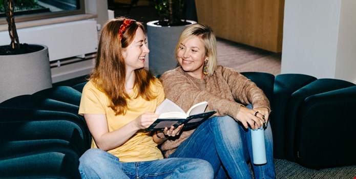 Två kvinnor sitter ner bredvid varandra på golvet och ler. Den ena visar sidor ur en bok för den andra.